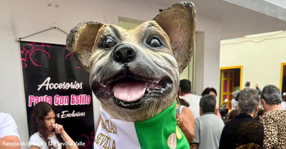 A statue of a smiling dog wearing a green bandana is displayed at a public event with people gathered in the background.