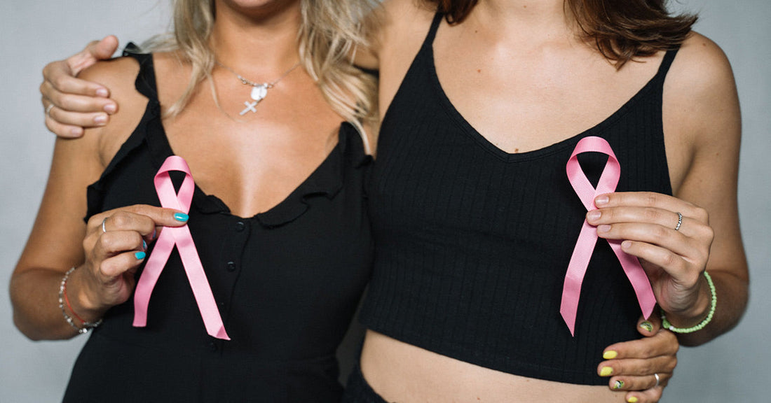 Two women stand side by side with arms around each other, each holding a pink awareness ribbon against their black tops.