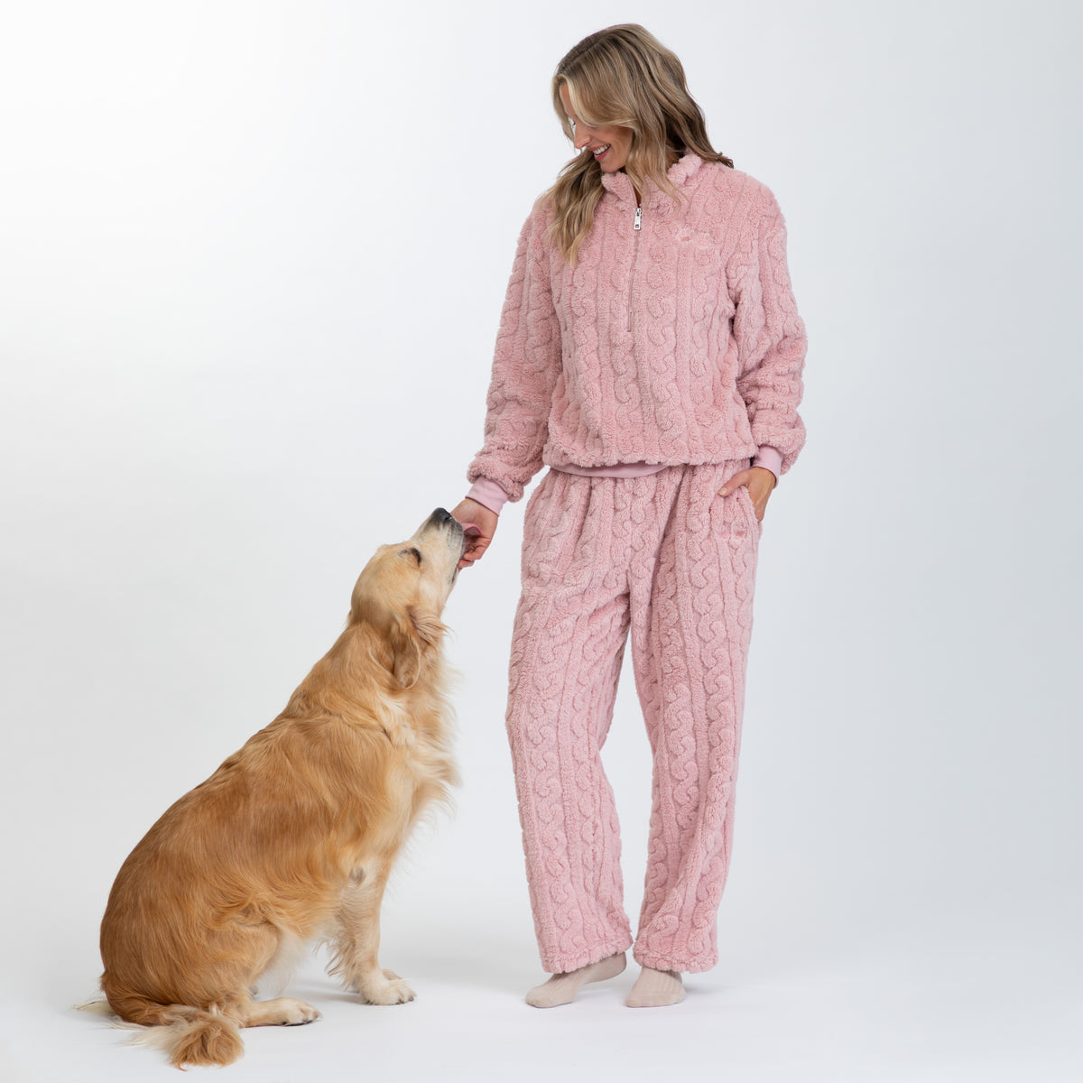A woman wearing a pink cable-knit fleece pajama set stands on a white studio background while a golden retriever dog reaches up to her hand. The set includes a matching zip-up top and pants.
