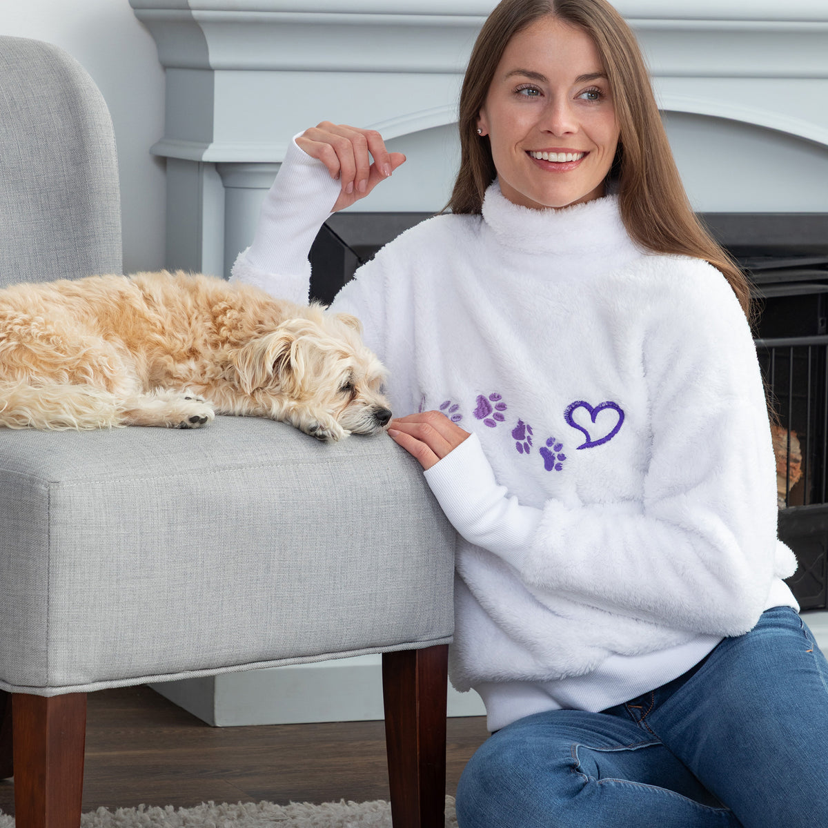 A woman in a white fuzzy mock neck sweater with purple paw prints and a heart sits next to a small fluffy dog resting on a gray armchair in a living room setting.