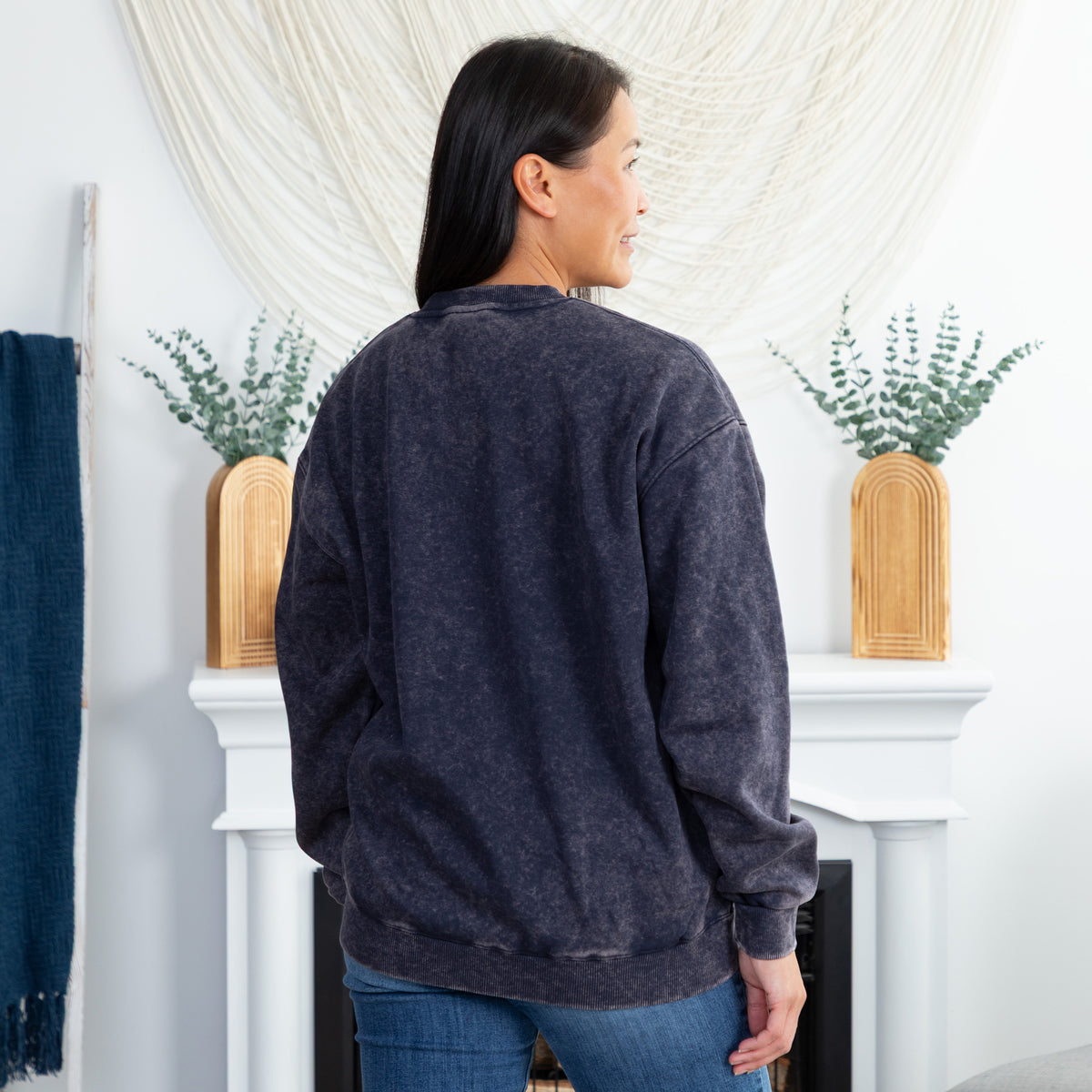 A woman viewed from behind wearing a dark washed, heathered charcoal crewneck sweatshirt and blue jeans, standing in front of a white fireplace mantel decorated with vases and greenery.