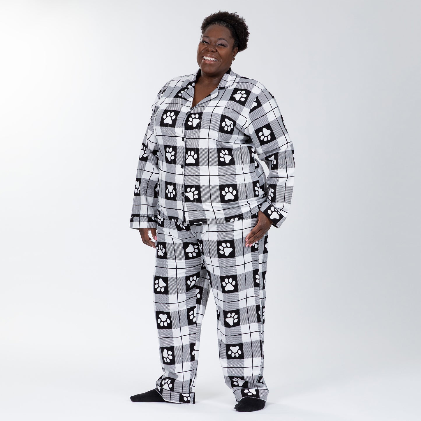A woman wearing a matching pajama set of white and black, plaid, button-up, long-sleeve top patterned with paw prints and pants of the same color and pattern. Hand in pocket of pajamas. Plus and extended size available.