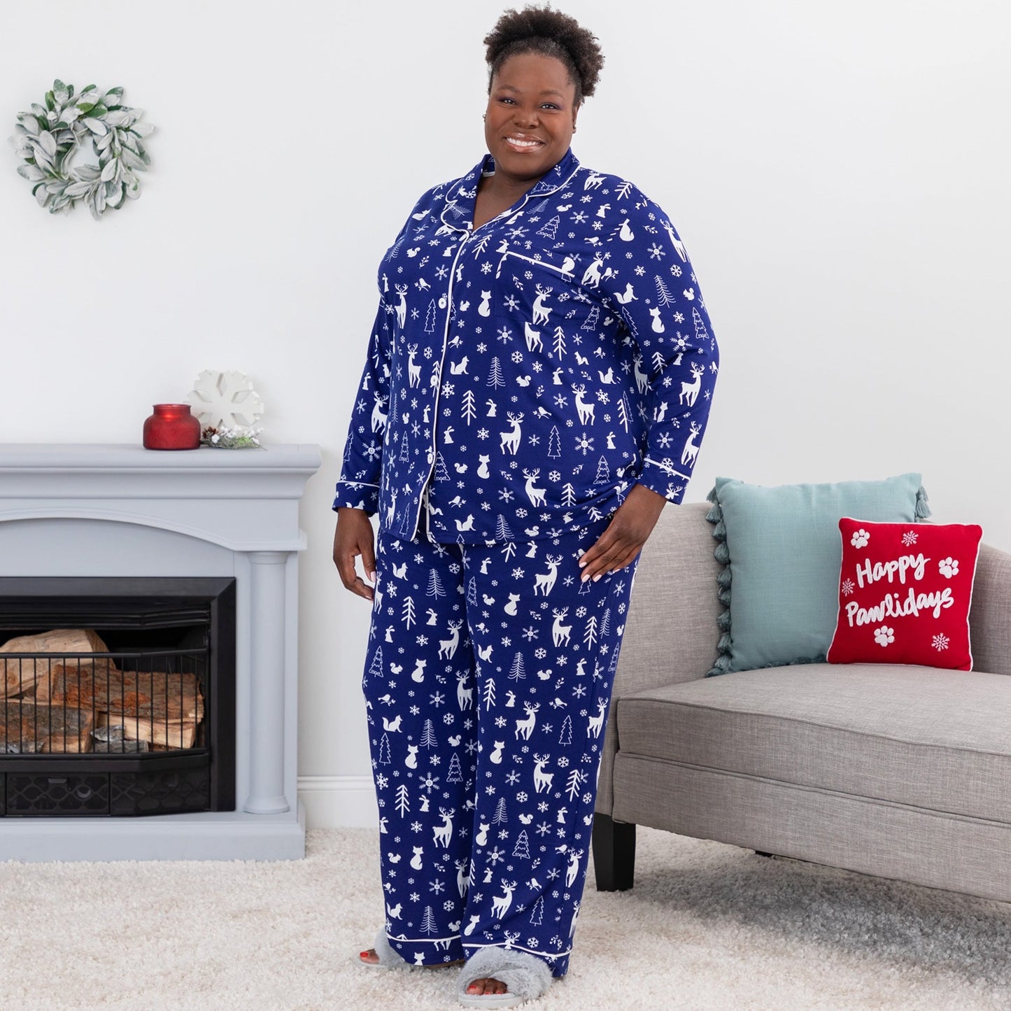 Woman standing in a bedroom wearing a soft pajama set, featuring a cute woodsy winter scene of forest animals surrounded by snowflakes. This deep blue PJ set has white design and white piping, it includes a button up top with chest pocket, paired with pants with an elastic waistband with a drawstring and pockets. Extended and plus size available.