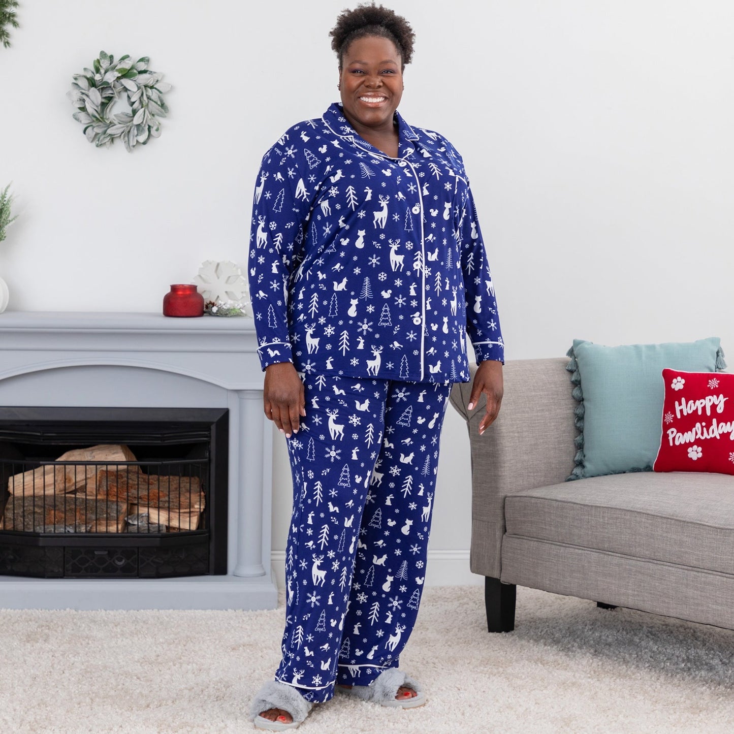 Woman standing in a bedroom wearing a soft pajama set, featuring a cute woodsy winter scene of forest animals surrounded by snowflakes. This deep blue PJ set has white design and white piping, it includes a button up top with chest pocket, paired with pants with an elastic waistband with a drawstring and pockets. Extended and plus size available.
