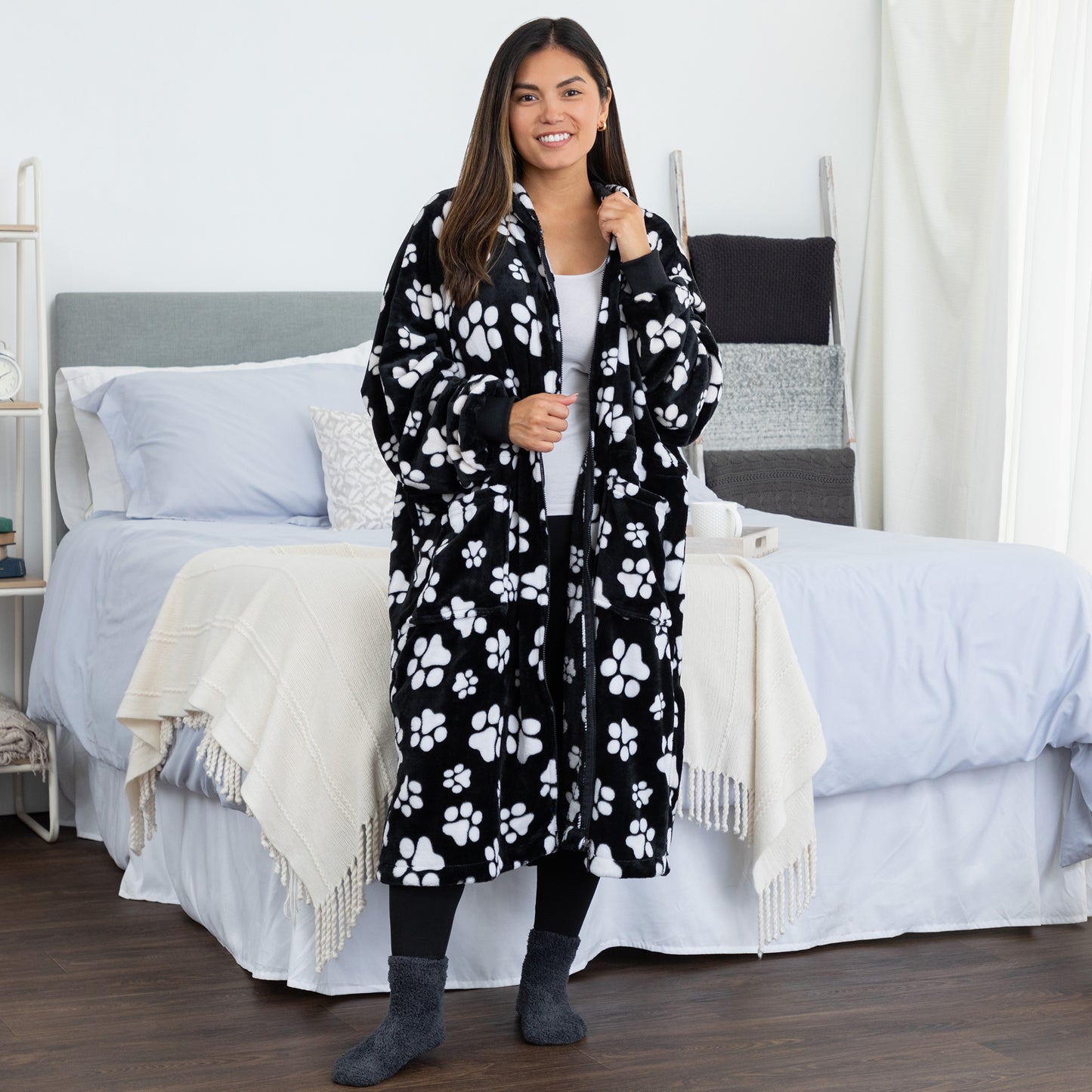 A woman wearing a black and white paw print patterned fleece blanket hoodie in a bedroom setting.