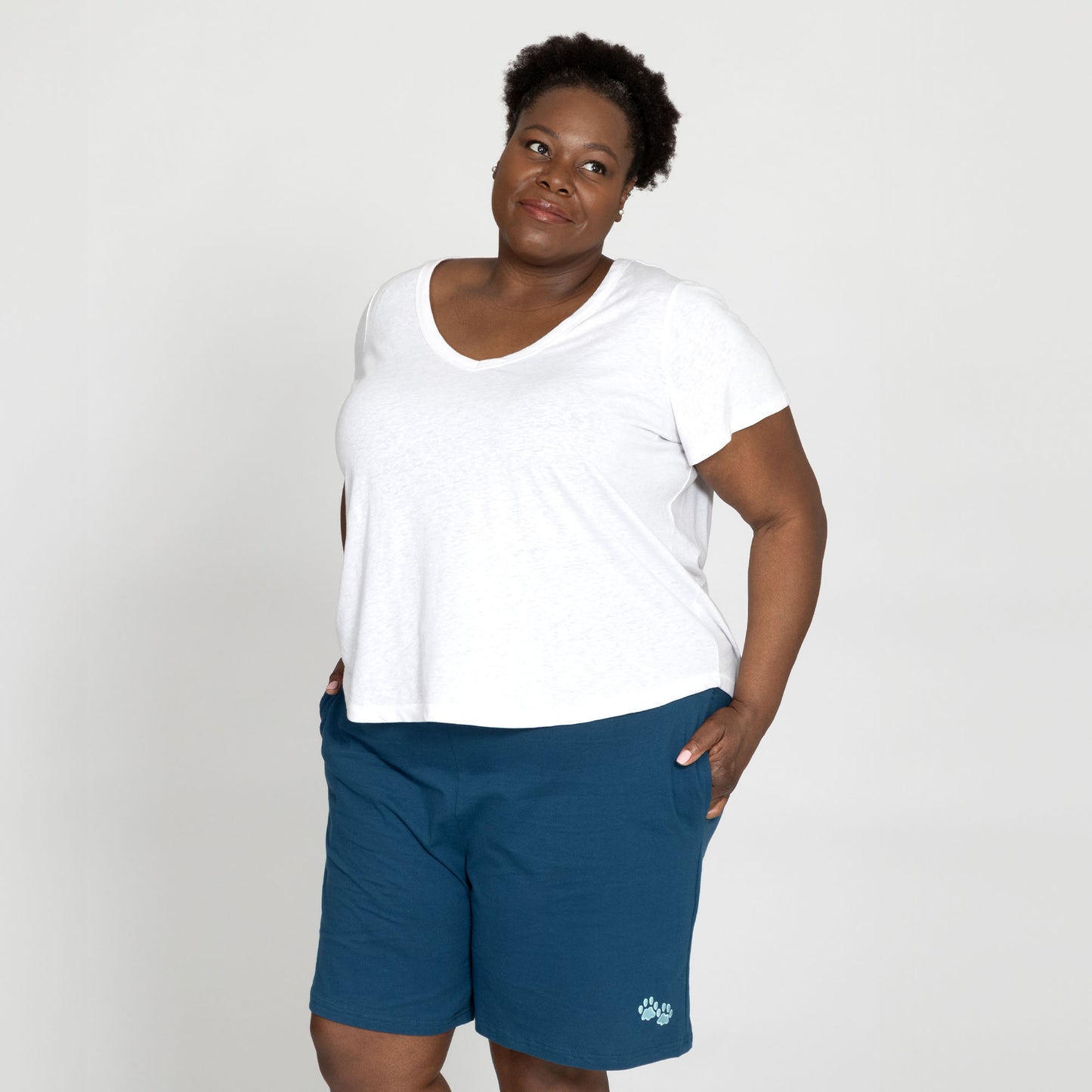 Woman wearing a white t-shirt and teal shorts, smiling against a neutral background.