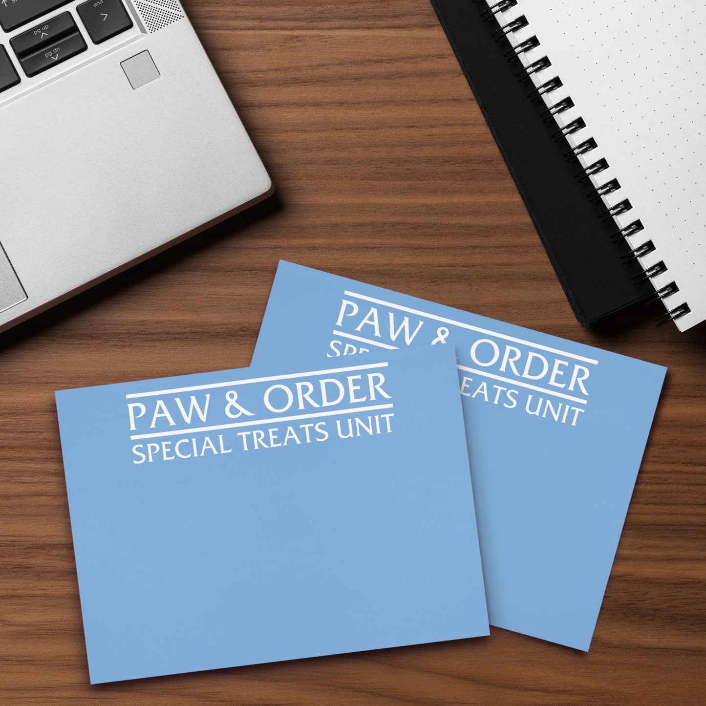 Closeup of Two blue cards with 'Paw & Order Special Treats Unit' text on a wooden surface with a laptop and notebook.