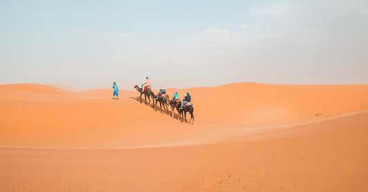 A caravan of camels with riders walking across golden sand dunes under a blue sky with scattered clouds.
