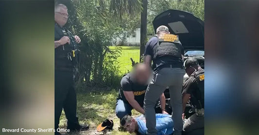 Police officers arrest a man who is pinned to the ground in a grassy, shaded area.