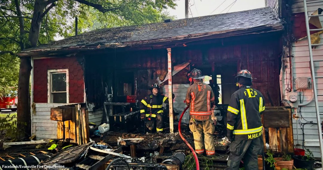 Three firefighters work amid charred debris outside a heavily fire-damaged red wooden structure, holding a hose.
