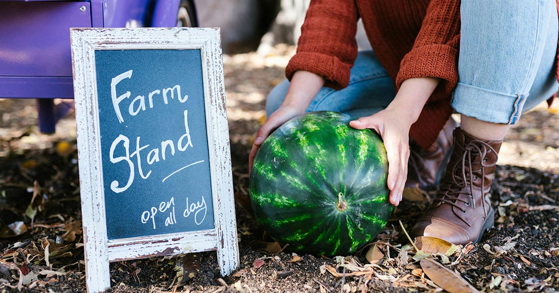 A closeup of handsgrasping a watermelon near a roadside farm stand sign..