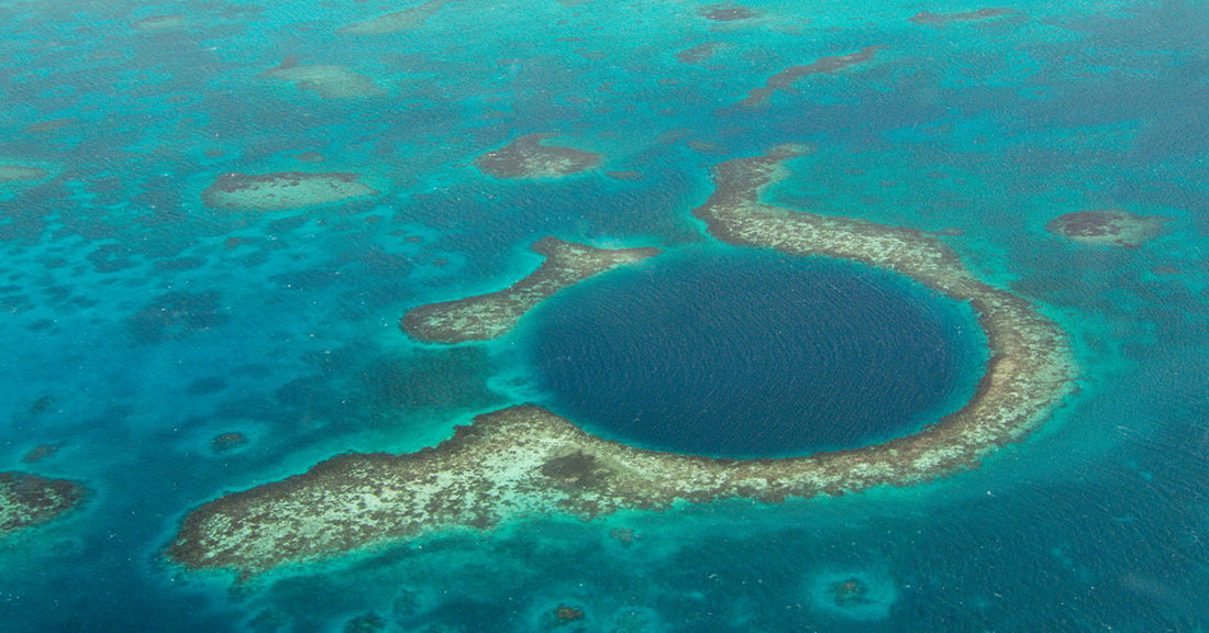 An aerial photograph of the Great Blue Hole in Belize, showing a perfect circular dark-blue sinkhole surrounded by turquoise shallows and scattered coral reefs.