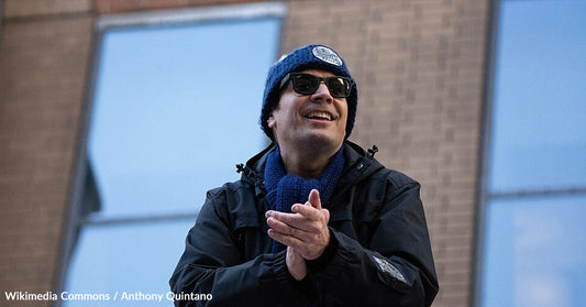 Jimmy Fallon wearing sunglasses, a winter hat, and scarf clapping outdoors near a brick building.