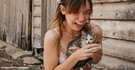 Smiling woman outdoors beside a weathered wooden wall cradles a tiny gray-and-white kitten while holding a small can of food.