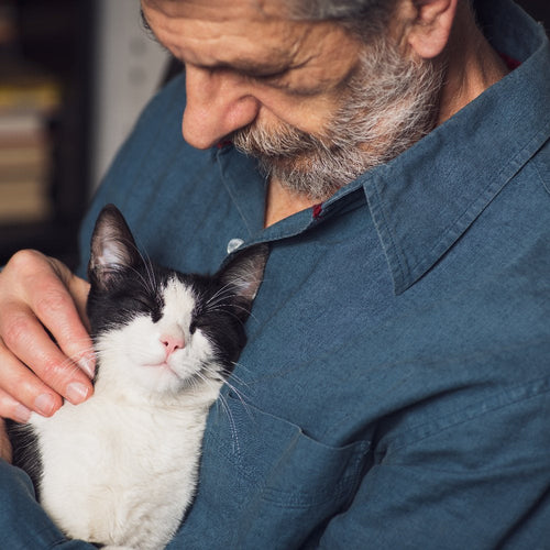 An older man with a gray beard gently cradles a black-and-white cat against his chest indoors.