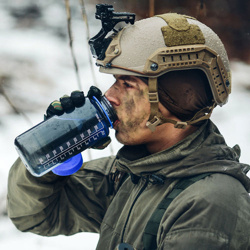 Soldier in cold-weather gear and helmet drinks from a large water bottle while standing outdoors in a snowy landscape.