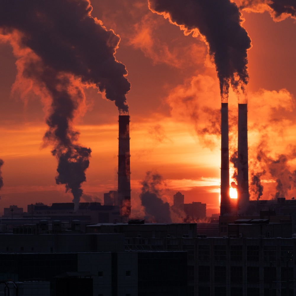 Several industrial smokestacks billow thick plumes of smoke against a vivid orange and red sunset sky over a city skyline.