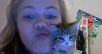 A young woman with blonde hair holds a small tabby kitten close to her cheek while making a kissing face indoors.