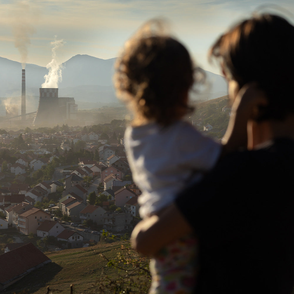 A woman holding a small child looks out over a town blanketed in haze, with a smoking industrial plant in the distance against mountain slopes.