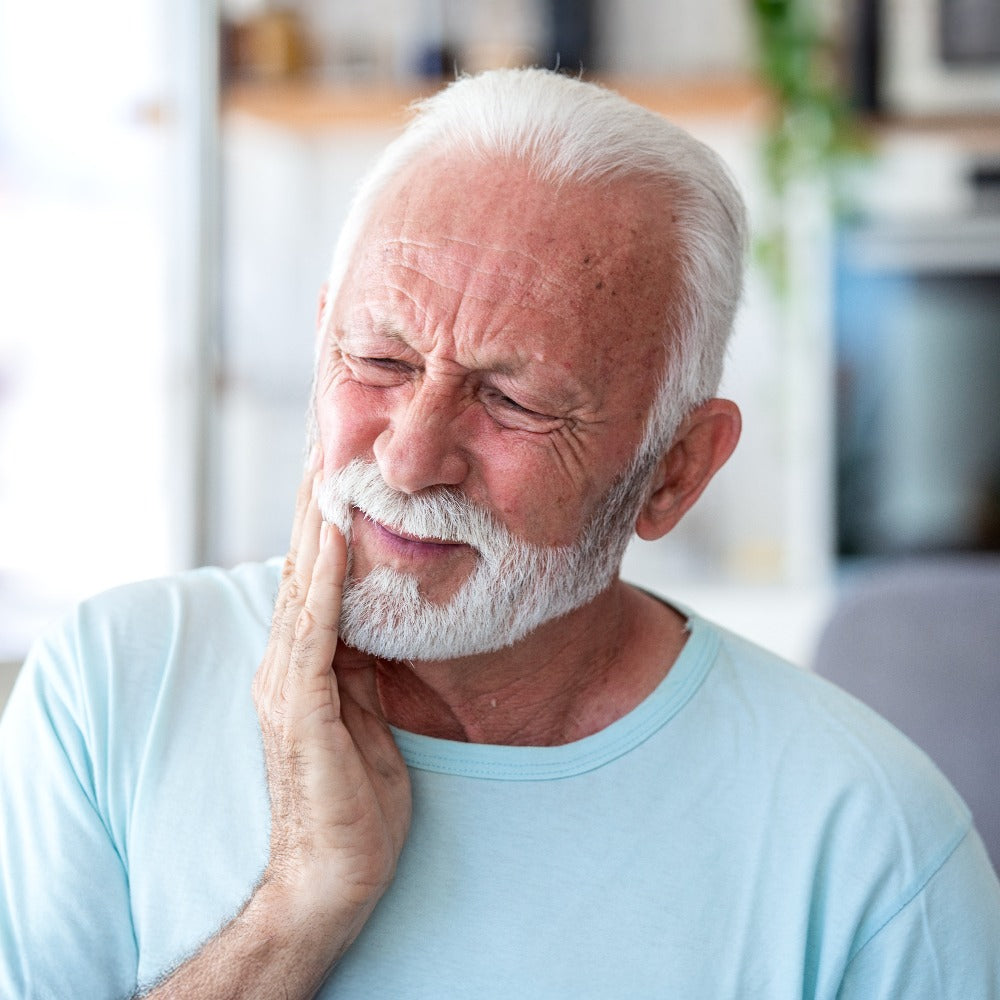 An elderly man with white hair and beard holds his jaw in pain while sitting indoors.