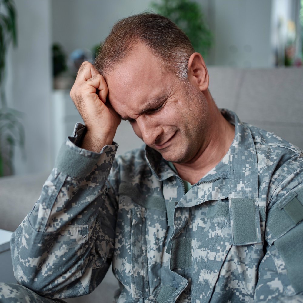 Man in military fatigues sitting with his eyes closed and his fist pressed against his forehead.