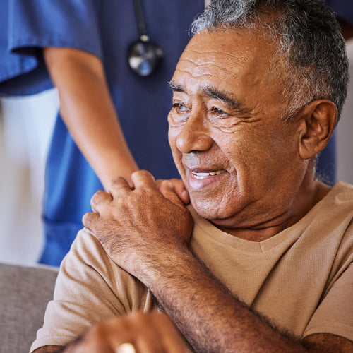 Close-up of an older man smiling while a caregiver in scrubs supports him with a hand on his shoulder.