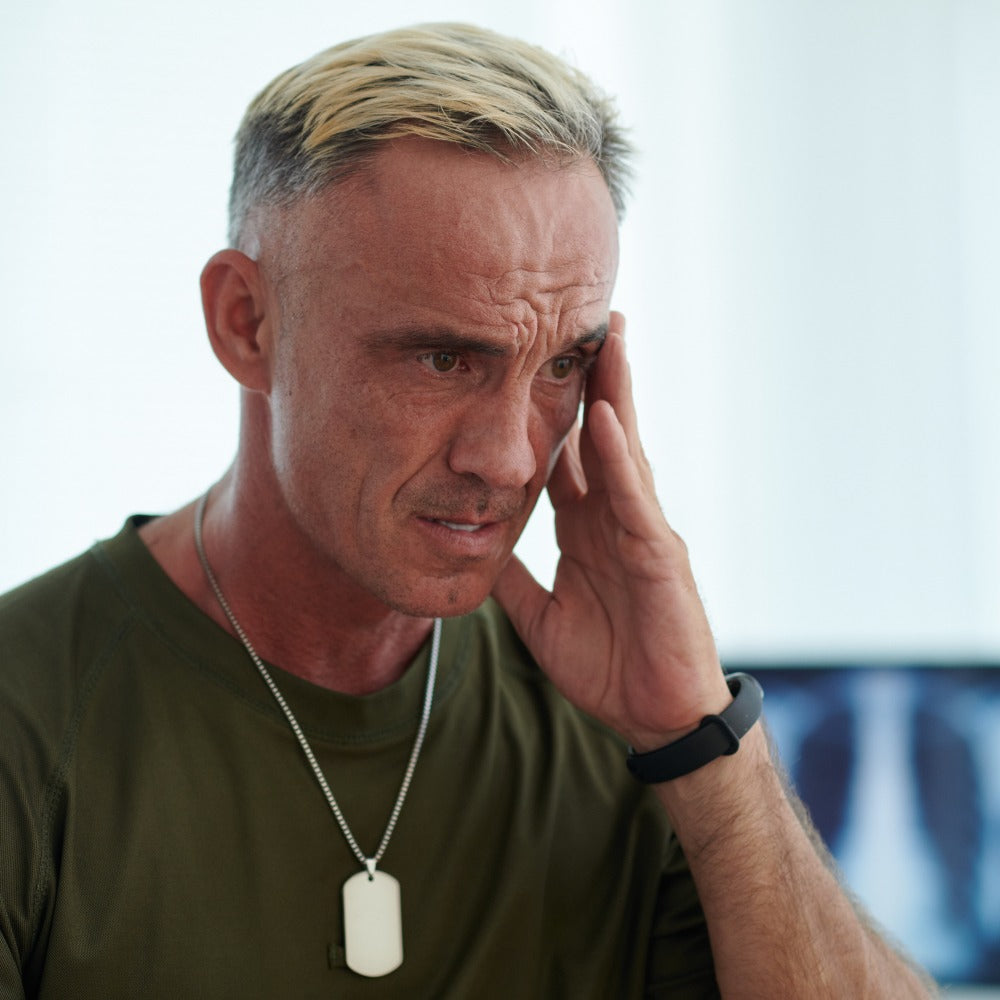 A middle-aged man wearing a military dog tag and olive green shirt holds his hand to his temple with a distressed expression, seated in a bright medical office.