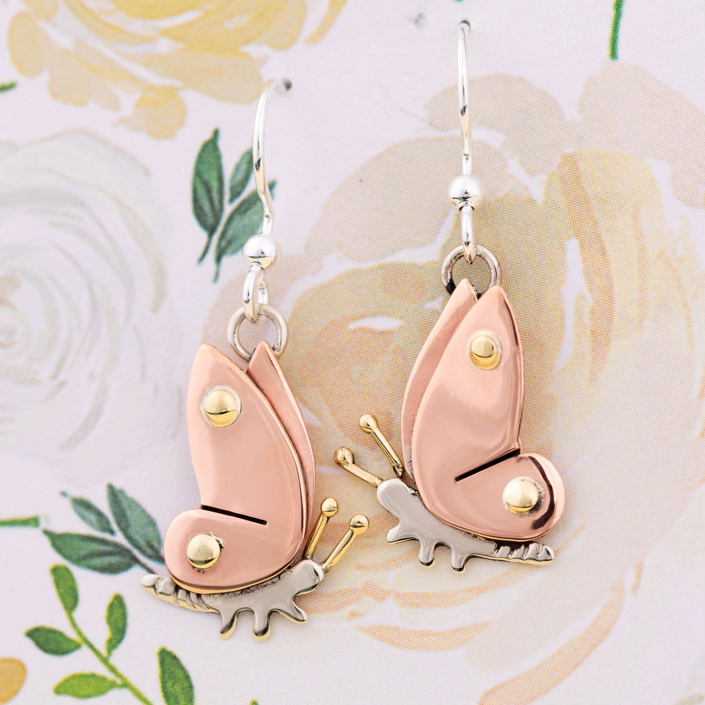 Butterfly-shaped earrings with copper, brass, and silver details on a floral background.