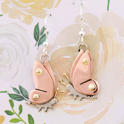 Butterfly-shaped earrings with copper, brass, and silver details on a floral background.