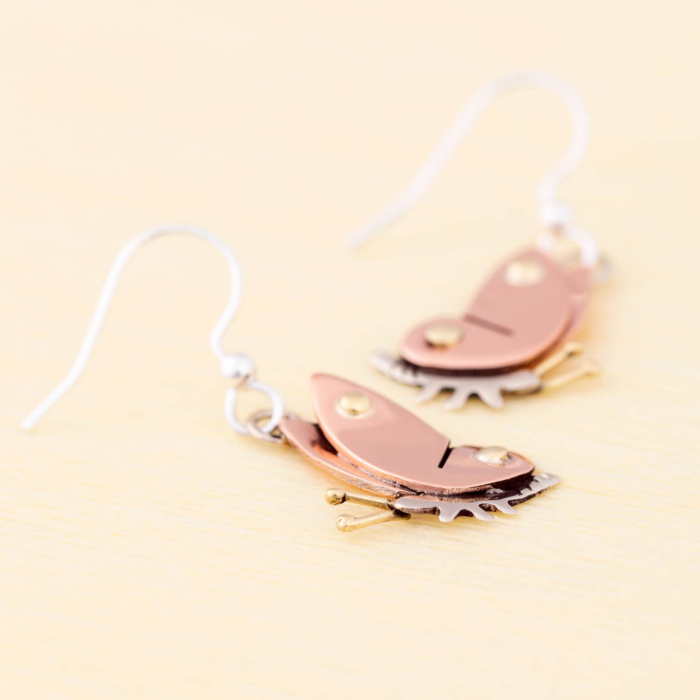 Butterfly-shaped earrings with copper, brass, and silver details on a light yellow background.