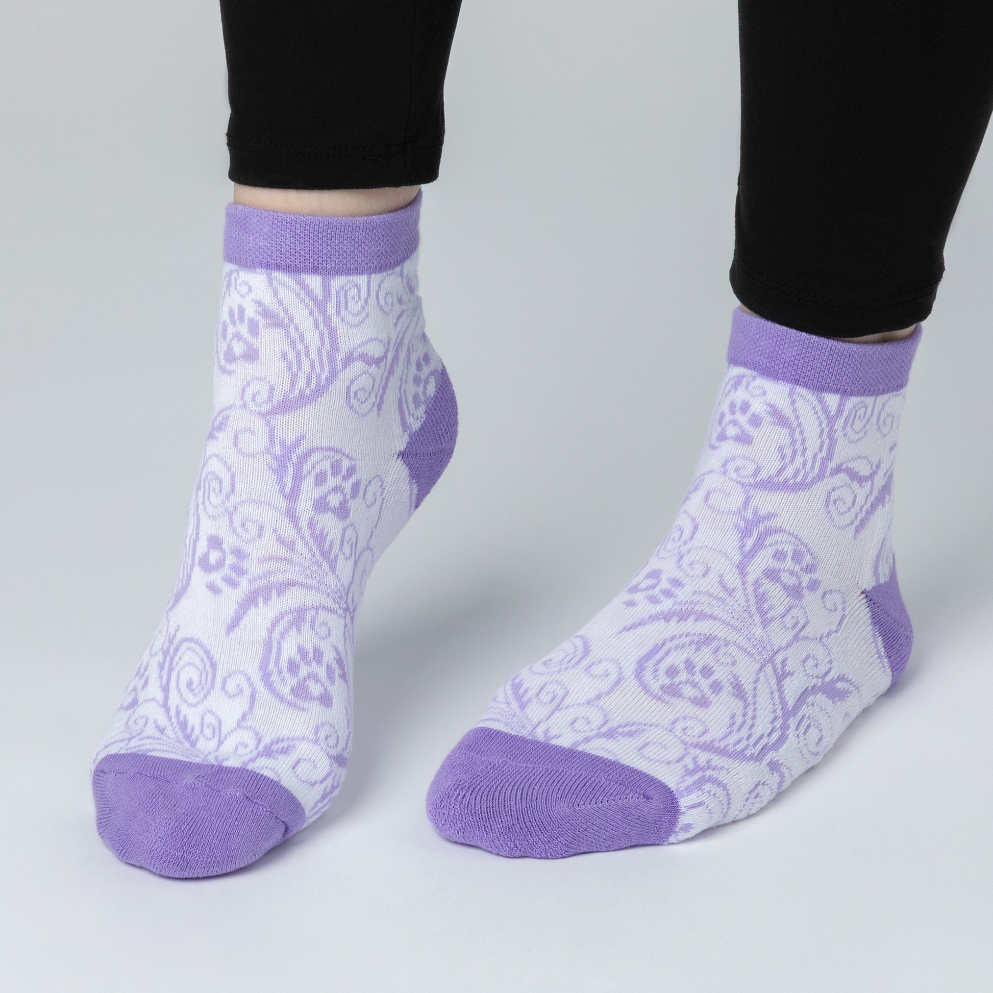 A person wearing white ankle length comfortable cotton-blend socks that are designed with a lavender paw print and swirl design. The cuff, heels and toes of the sock are a plain lavender color.  