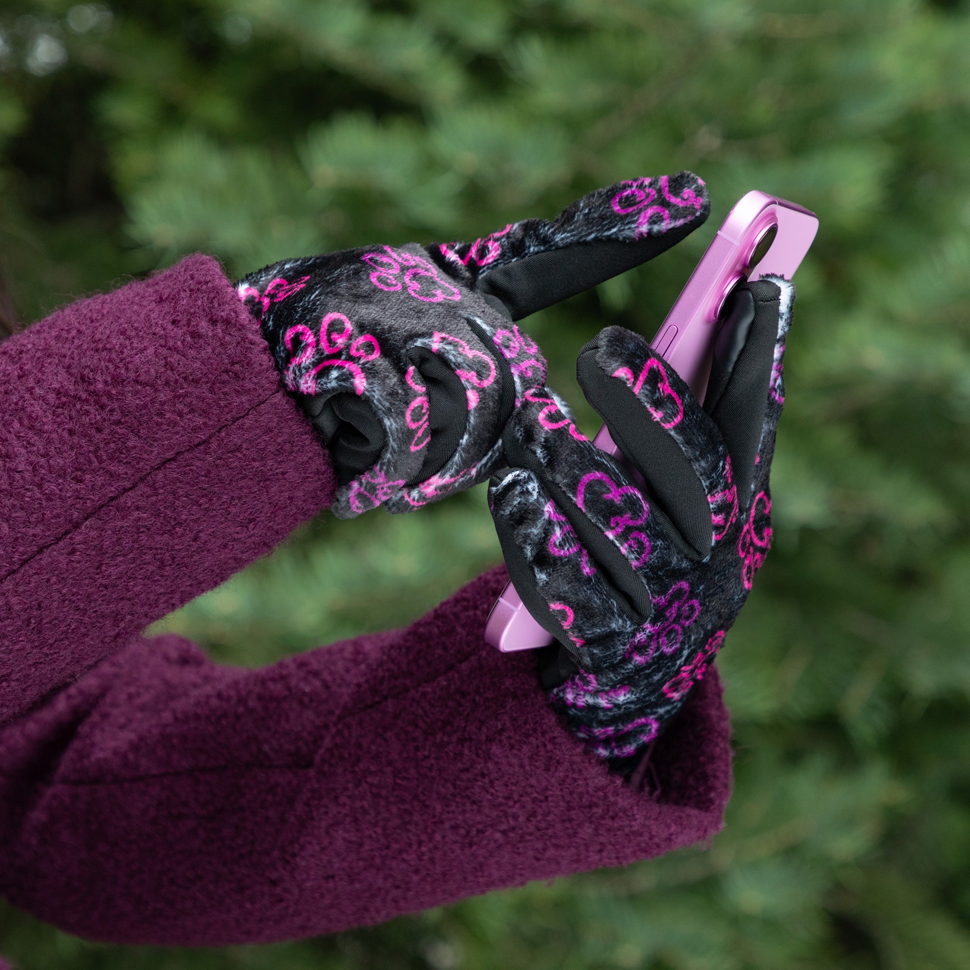A woman, outside, wearing a coat and a pair of touchscreen technology fingertips warm black fleece gloves adorned with prink and violet elegant paw prints. She uses the touchscreen technology fingertip with her thumb and index finger on her touchscreen phone.