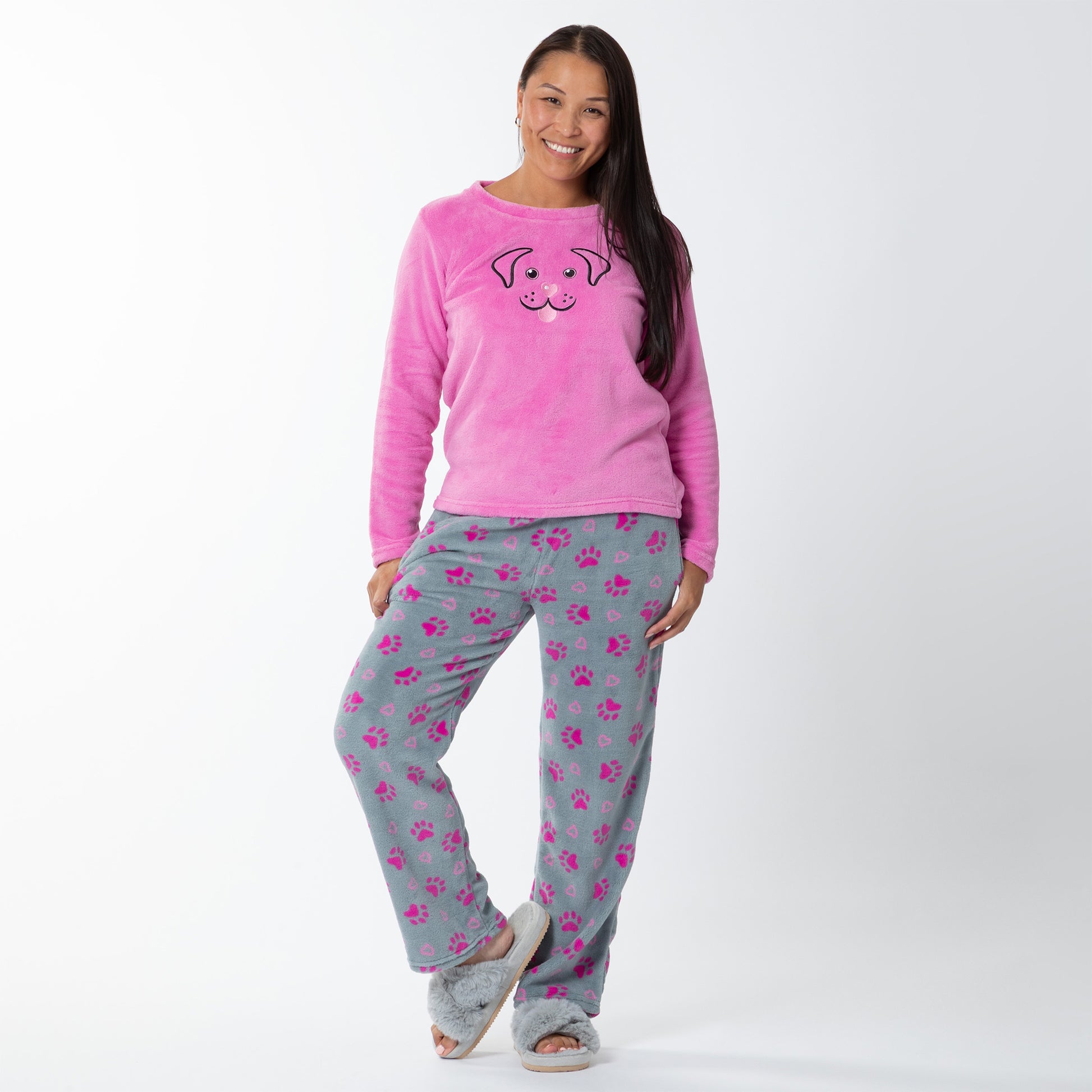 A woman wearing a pink long-sleeve top with an embroidered dog face design and gray pants with pink paw prints and hearts pattern. Standing against white background Design a pink cat in super cozy fabric, showing an Elastic waistband with a drawstring, two side pockets