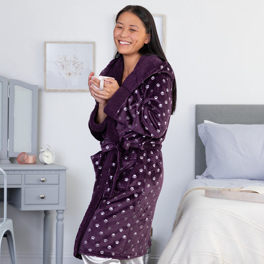 A woman wearing a purple hooded robe with a silver paw print pattern standing in a bedroom, holding a coffee cup.