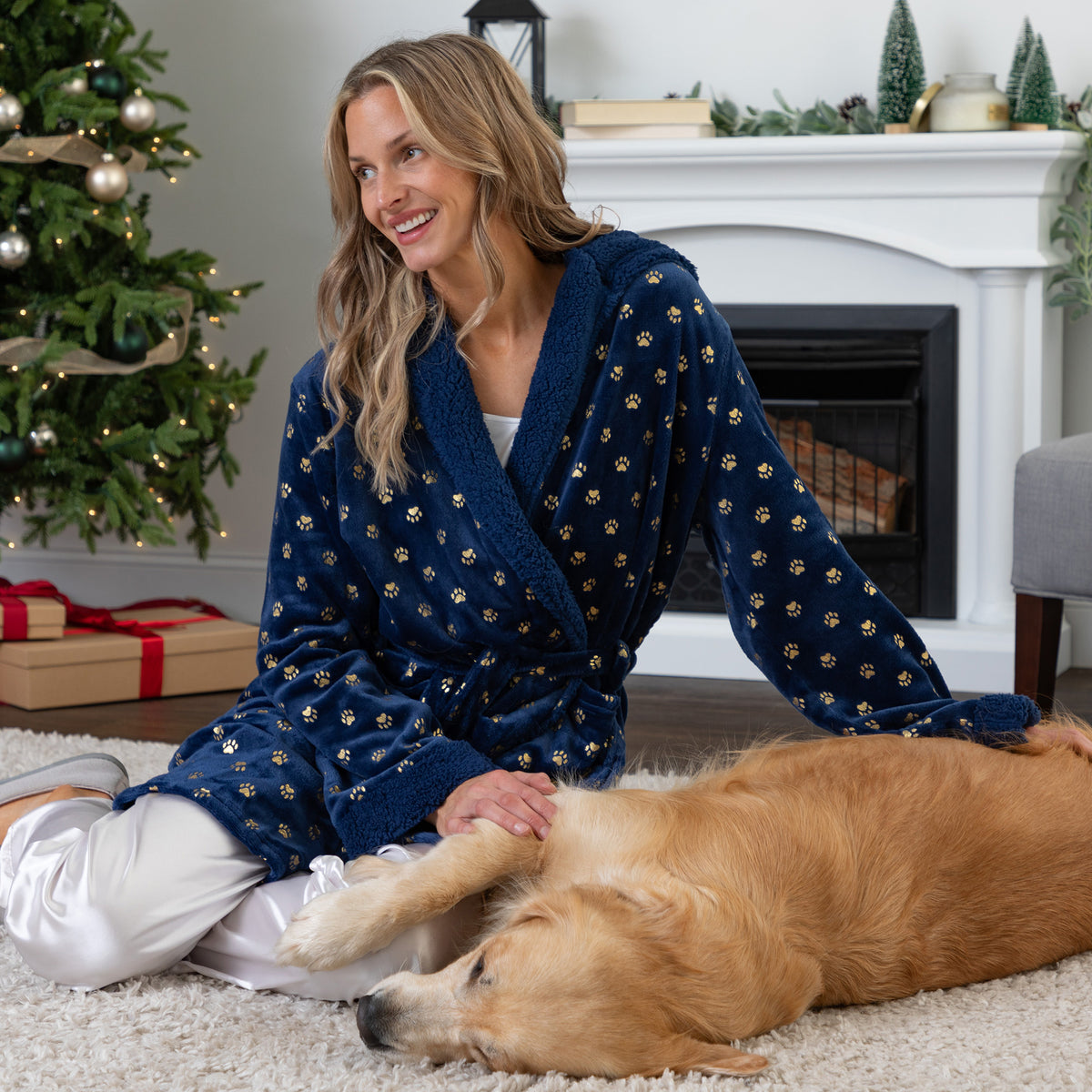 A woman wearing a navy blue plush robe with small gold anchor patterns sits on a white carpet in a living room, petting a golden retriever dog. A decorated Christmas tree and fireplace are visible in the background.