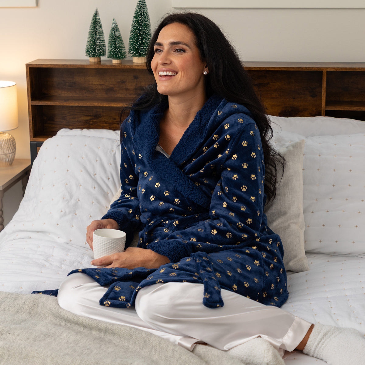A woman sitting on a bed wearing a navy blue plush robe with small gold paw prints and white pajama pants, holding a white mug in a cozy bedroom setting.