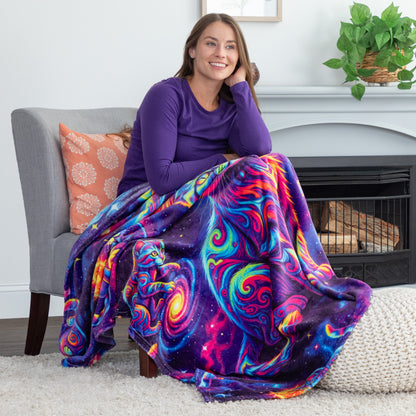 A woman sitting in a gray armchair in a living room, wrapped in a vibrant, multicolored galaxy swirl blanket. The blanket features a psychedelic pattern of purple, blue, pink, yellow, and green with starry details.