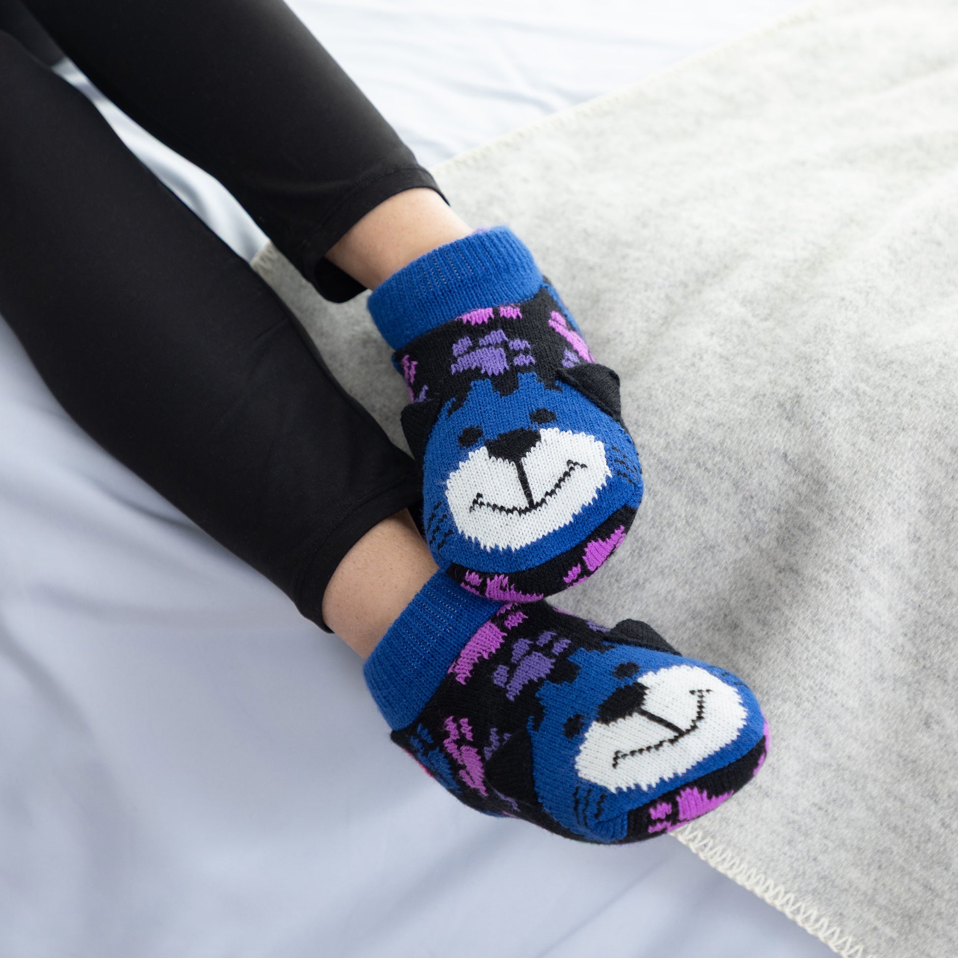 A person wearing black leggings and blue knit booties with a cat face design and purple pattern details, resting on a light gray blanket.