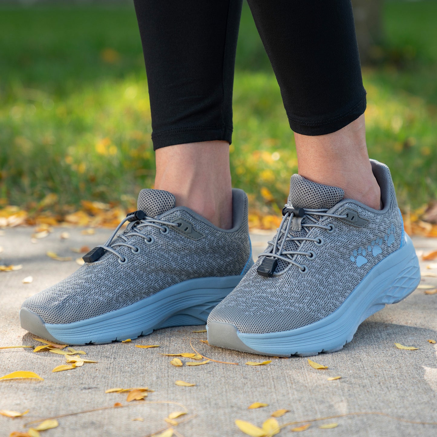 Gray sneakers with blue soles and three blue paw prints on the side, worn by a person on a leafy ground.