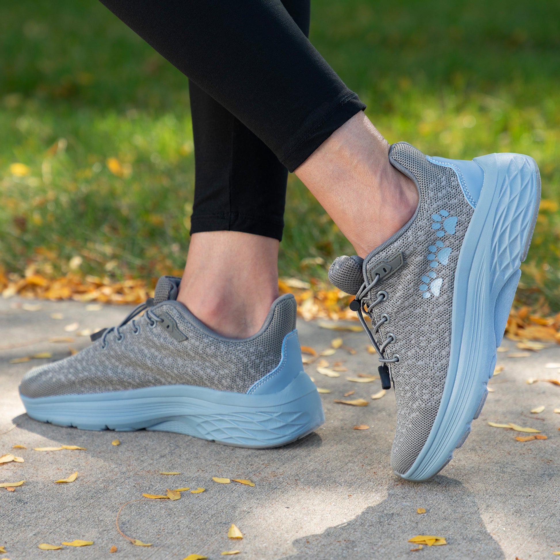 Gray and blue running shoes with bungee laces and three blue paw prints on each side, worn outdoors on a paved path with grass and leaves in the background.