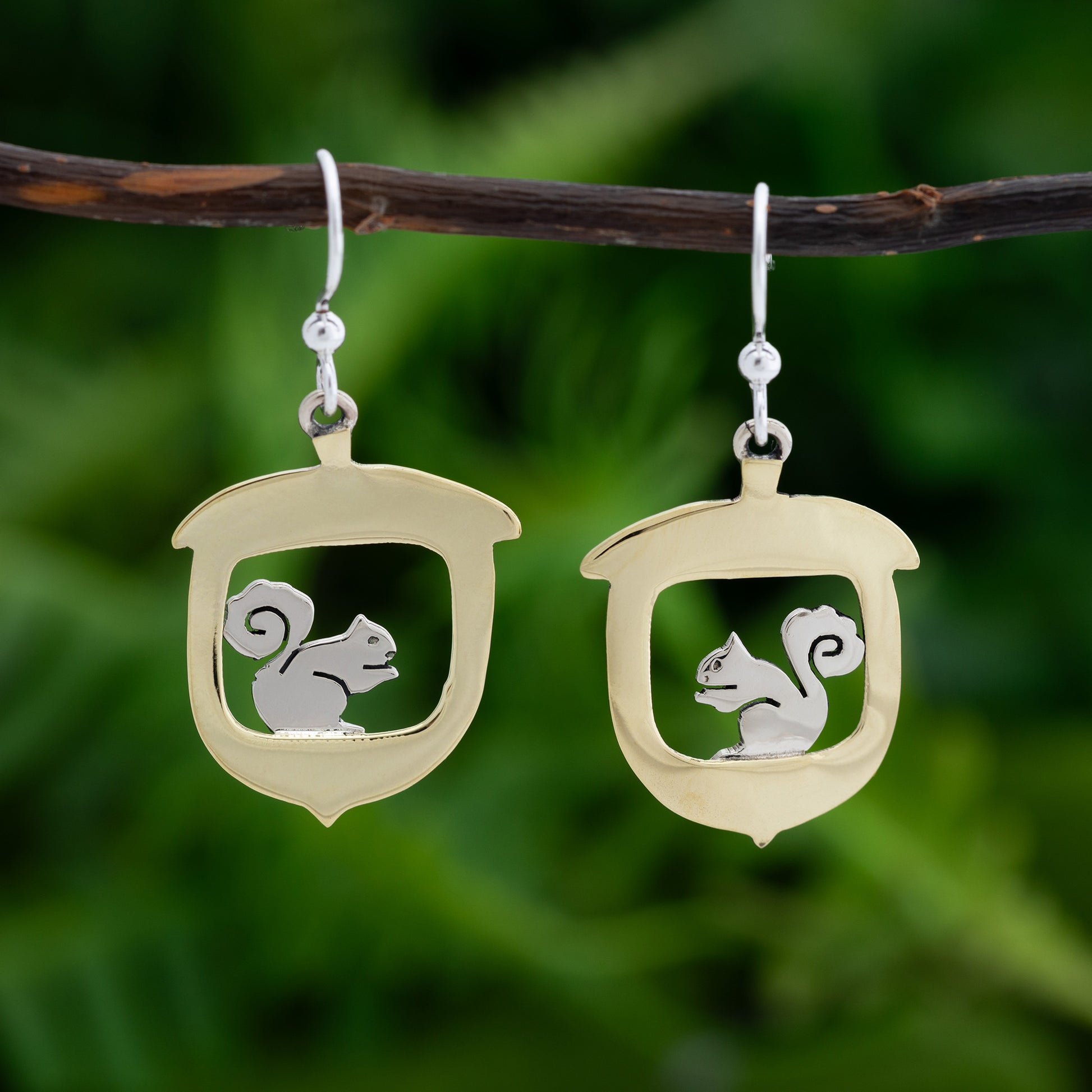 Brass earrings with a squirrel design on a branch against a green background.