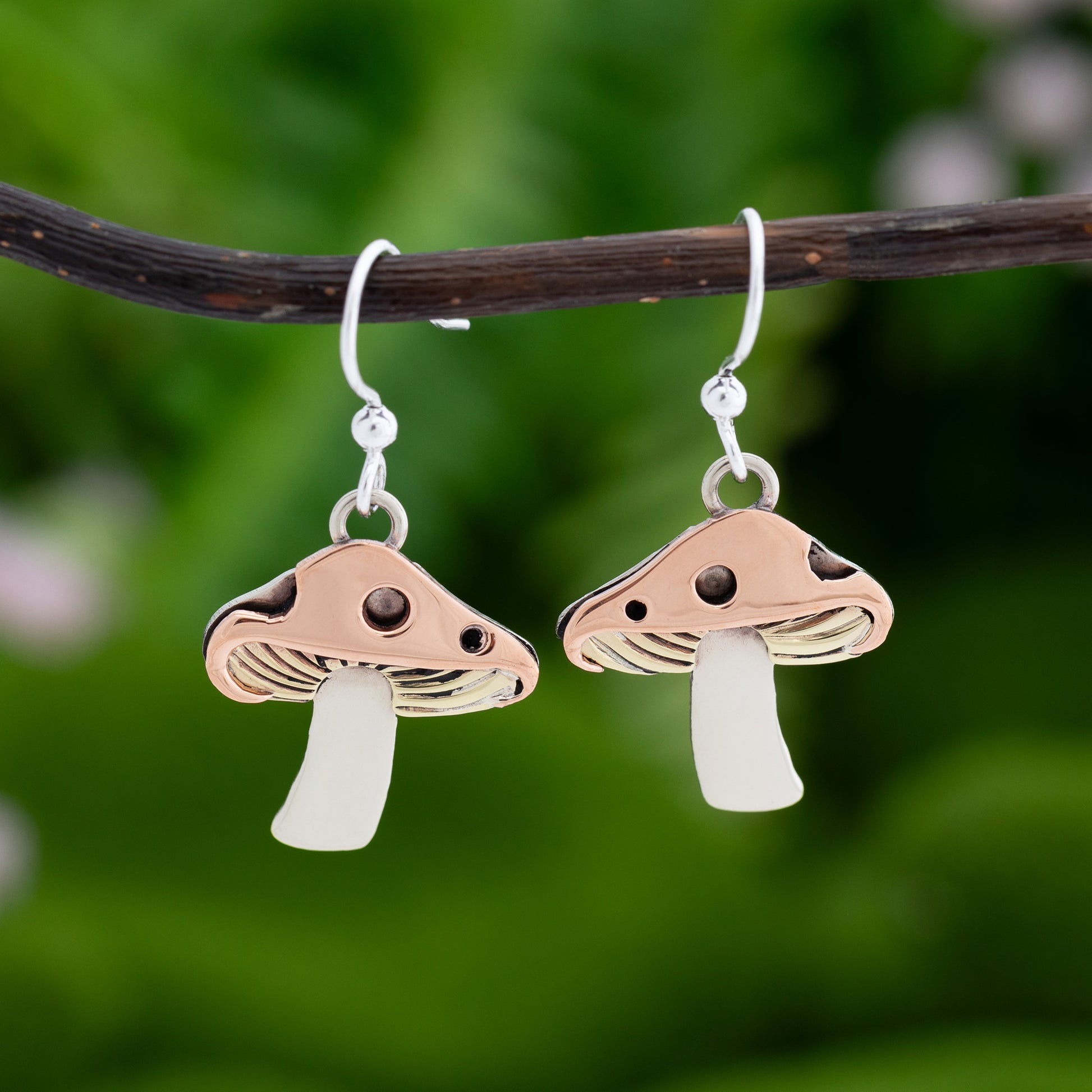 Mushroom-shaped earrings with copper, brass, and silver, hanging on a branch with a natural green background.