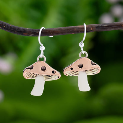 Mushroom-shaped earrings with copper, brass, and silver, hanging on a branch with a natural green background.