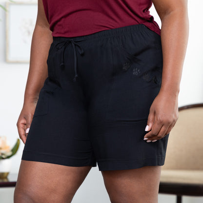 Person wearing black shorts with two embroidered paws on the pocket, in front of a blurred indoor background.