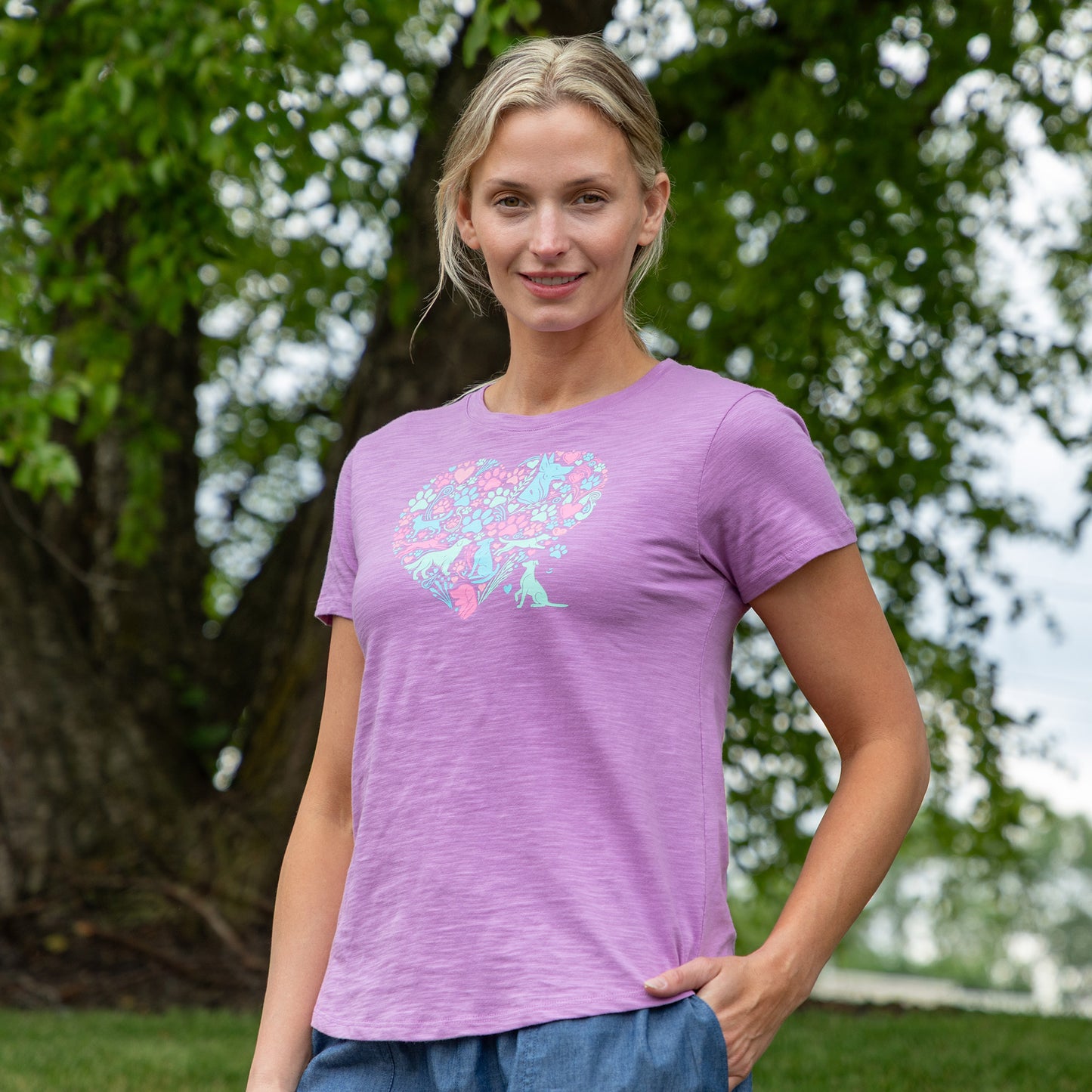 A woman wearing a heathered purple short-sleeve t-shirt with a pastel floral graphic on the front, standing outdoors in front of a green tree.