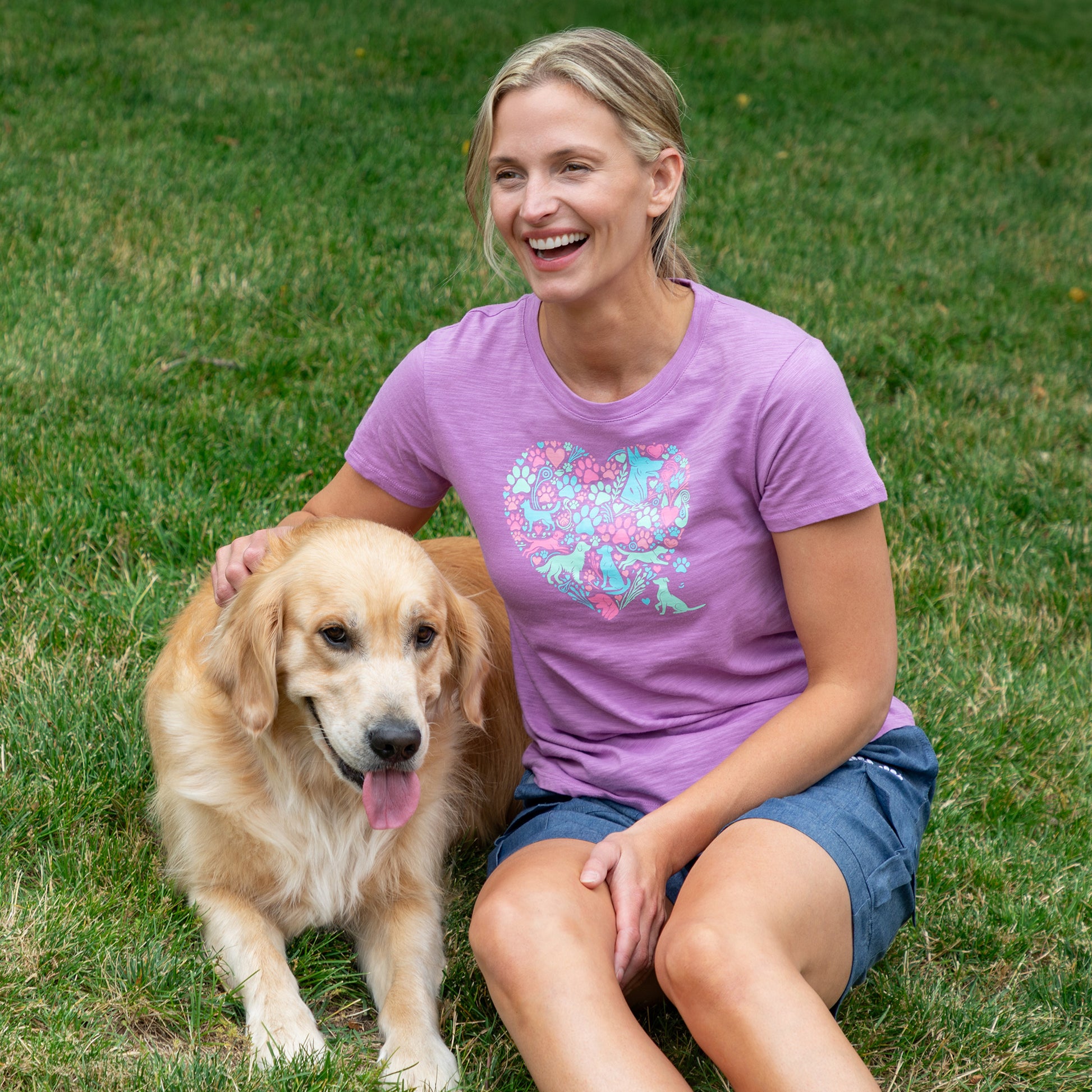 A woman sitting on green grass next to a golden retriever dog, wearing a purple short-sleeve t-shirt with a colorful floral heart graphic on the front.