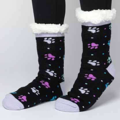 model wearing Pair of black with purple and blue polka dot paws Sherpa Fleece Lined Paw Print Cabin Slipper Socks with sherpa trim on a light gray background polka dot paws 