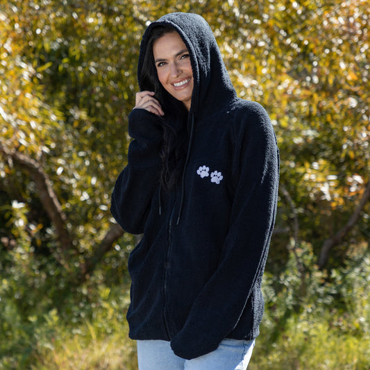 Woman standing outside with trees in the background wearing a black hooded sherpa fleece zip up jacket, with two embroidered paws on the chest. 