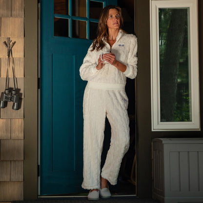 Woman leaning in doorway of cozy cabin. She is wearing a white, fleece, cable-knit, quarter-zip, long-sleeve shirt with two light blue paw prints embroidered beneath the lapel. Her pants are part of the matching set and are also white with a cable-knit pattern and two light blue paw prints embroidered on the lefthand pocket. 