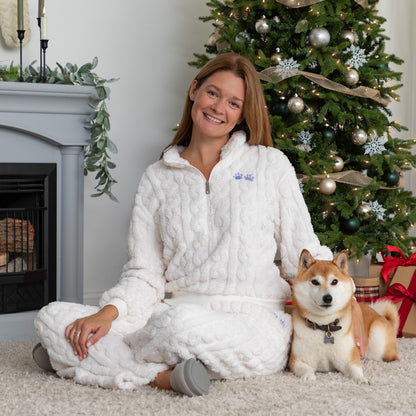 Woman sitting on the floor in cozy holiday living room setting with her arm around her pet dog. She is wearing a white, fleece, cable-knit, quarter-zip, long-sleeve shirt with two light blue paw prints embroidered beneath the lapel. Her pants are part of the matching set and are also white with a cable-knit pattern and two light blue paw prints embroidered on the lefthand pocket. 